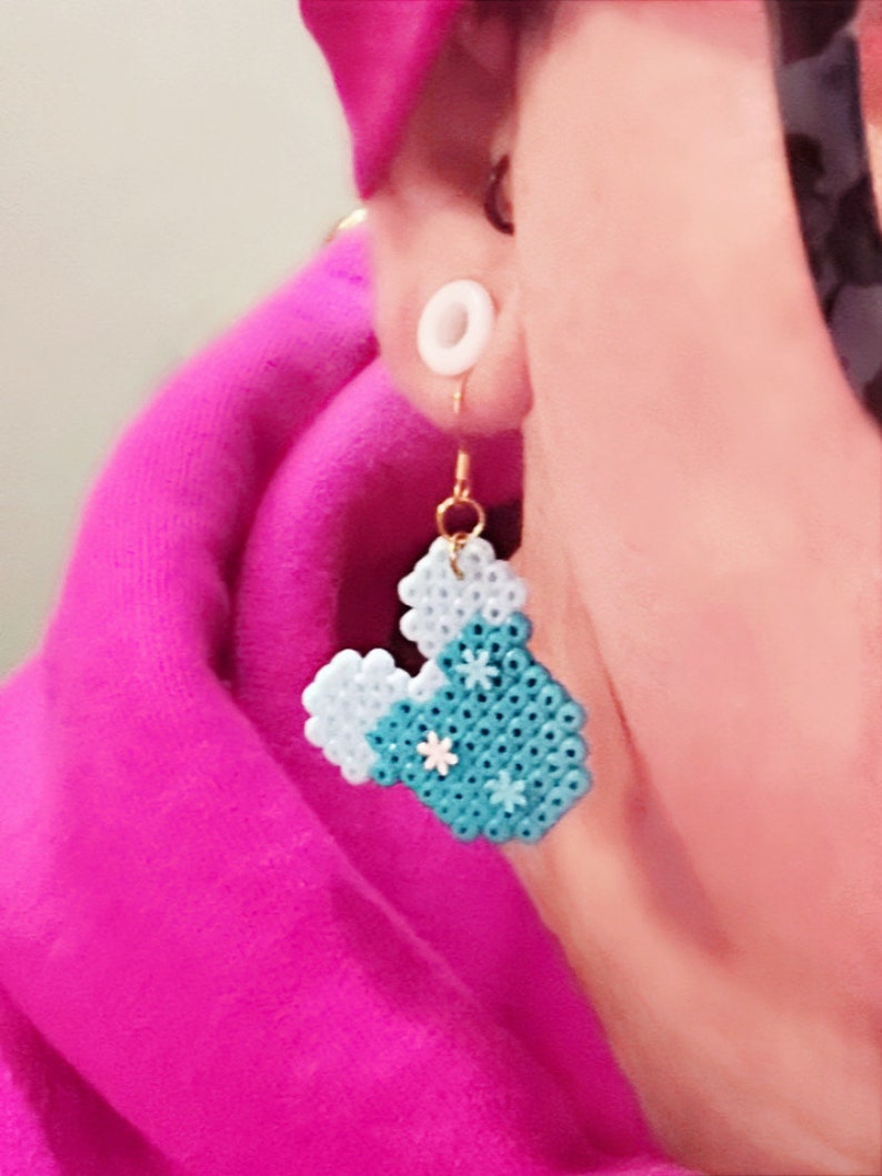 Puede incluir: Un par de pendientes en forma de coraz&oacute;n azul y blanco con flores blancas. Los pendientes est&aacute;n hechos de peque&ntilde;as cuentas y cuelgan de ganchos dorados.