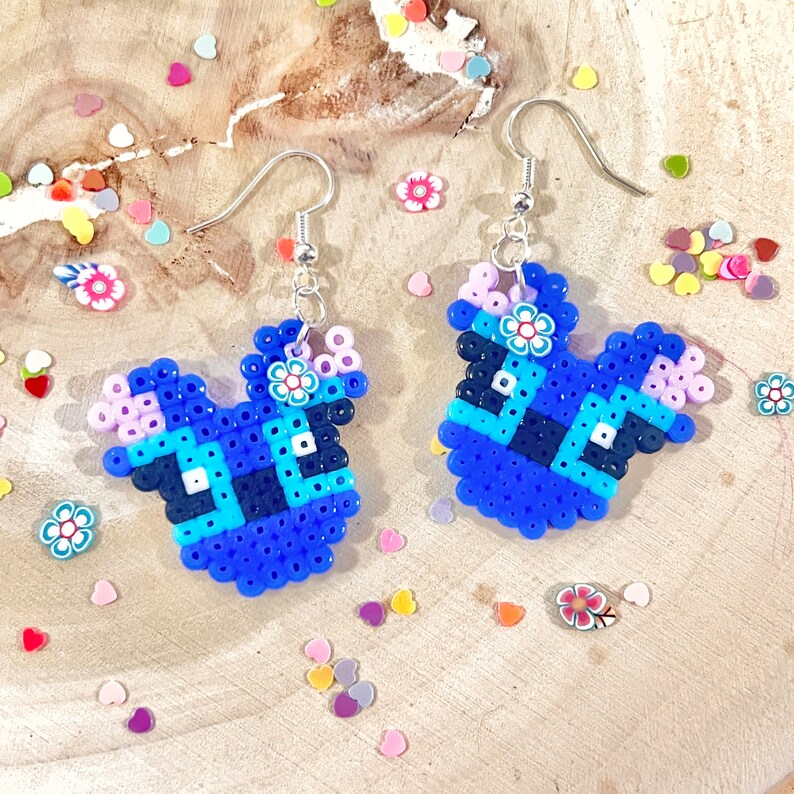 Puede incluir: Un par de pendientes de cuentas azules, rosas y blancas con forma de personaje de dibujos animados. Los pendientes cuelgan de ganchos plateados.