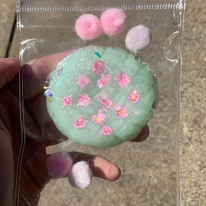 May include: A round, light mint green wax melt with a waffle-like grid pattern, embedded with small pink flower-shaped wax inserts and iridescent glitter, packaged in a clear plastic pouch with four small, fuzzy pink pom-poms inside.
