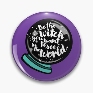 May include: A round pin featuring a purple background and a black crystal ball illustration. The crystal ball has a teal base and white text that reads, Be the witch you want to see in the world, surrounded by small white star and dot accents.