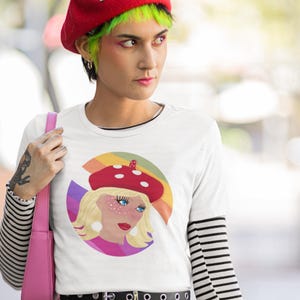 Op de afbeelding: Een wit T-shirt met korte mouwen en een ronde grafische print van een blond persoon met een rode baret met paddenstoelenpatroon. De achtergrond van de afbeelding bevat een regenboogkleurig boogontwerp met paarse, oranje en gele strepen.