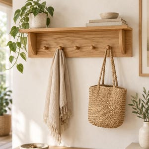 May include: A light-toned wooden wall-mounted shelf featuring four rounded pegs for hanging items. The shelf holds a potted green plant, a small bowl, and stacked books. A beige fringed textile and a woven straw tote bag hang from the pegs.