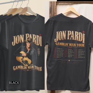 May include: A black t-shirt featuring a graphic of Jon Pardi with a guitar on the front and the Gamblin' Man Tour schedule with city names and dates printed on the back. The text includes Jon Pardi, Gamblin' Man Tour, and various locations.