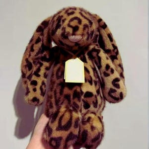 May include: A plush stuffed animal in the shape of a rabbit with long ears, covered in a brown and black leopard print pattern. The toy features a soft, fuzzy texture and has a blank, light yellow rectangular tag hanging from its neck area.