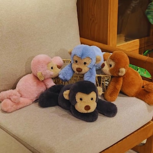 May include: A collection of four plush monkey toys in pink, light blue, dark gray, and brown. Each stuffed animal features a soft, fuzzy texture, a light tan face with a brown nose, and small rounded ears. The monkeys are arranged together.