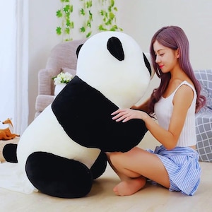 May include: A large, plush panda toy featuring a round white head with black patches around the eyes and black ears. The body is primarily white with large black patches on the arms and legs, crafted from soft, fuzzy material in a seated position.