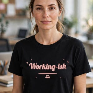 May include: A black short-sleeved t-shirt featuring the text "Working-ish" printed in a light pink cursive font. Below the text is a small, simple graphic of a computer monitor, accented by four small four-pointed star shapes surrounding the text.