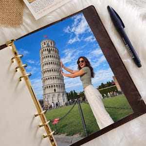 May include: A brown leather-bound planner with gold-tone metal rings containing a photograph of a person posing with the Leaning Tower of Pisa. A dark blue pen rests on the side, and a card with the text Always worth it is visible.