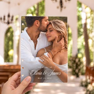 May include: A rectangular paper insert with six punched holes along the left edge, featuring a photograph of a couple embracing. The text reads THE STORY OF Olivia & James MAY 15, 2027. The image shows a person kissing another person on the forehead.