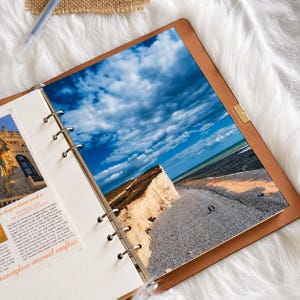 May include: A tan leather ring binder planner featuring a large photograph of white chalk cliffs along a pebble beach under a bright blue sky with clouds. The left page displays a smaller image of a building and printed text in orange and black.