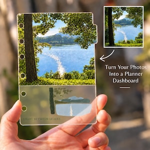 May include: A rectangular clear plastic planner dashboard featuring a printed landscape photograph of a lake, trees, and sunlight. The lower section includes a stylized graphic of the same scene with the text "LIGHT BETWEEN LEAVES" printed below.