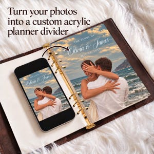 May include: A custom acrylic planner divider featuring a photograph of a couple embracing on a beach at sunset. The text reads: THE STORY OF Olivia & James MAY 15, 2027. The divider is shown inside a ring-bound planner alongside a smartphone.