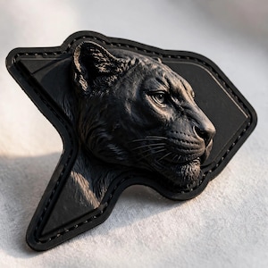 May include: A black patch featuring a three-dimensional relief of a panther head in profile. The panther is depicted with detailed fur texture, pointed ears, and focused eyes. The patch has a stitched border and an irregular, angular shape.