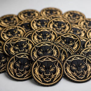 May include: A collection of circular embroidered patches featuring a black panther head design with gold thread detailing. Each patch has a serrated edge and displays a stylized feline face with sharp eyes and whiskers, arranged in a dense cluster.