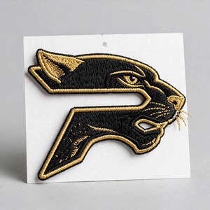 May include: An embroidered patch featuring a stylized black panther head profile integrated into a letter P shape. The design is outlined and detailed with metallic gold thread against a black background, presented on a white square backing card.