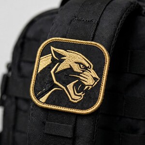May include: A square-shaped embroidered patch featuring a gold-colored panther head in profile with an open mouth, set against a black background with a gold border, attached to the black fabric strap of a tactical backpack.