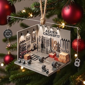 May include: A miniature gym diorama Christmas ornament featuring a gray floor and walls with the text ALEX'S CROSSFIT. The scene includes a barbell, kettlebells, a squat rack, wooden boxes, and wall-mounted gym equipment, suspended by a twine loop.