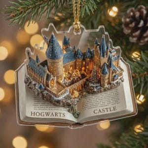 May include: A clear, book-shaped Christmas ornament featuring a detailed, three-dimensional model of a castle with blue-roofed towers and warm interior lighting. The open book pages display the printed text HOGWARTS CASTLE at the bottom.