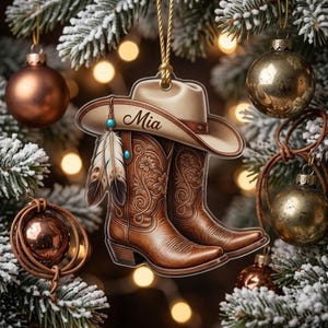May include: A flat, hanging Christmas ornament featuring a beige cowboy hat with the name Mia printed on the brim, paired with a pair of brown leather cowboy boots decorated with floral patterns and two hanging feathers with turquoise accents.