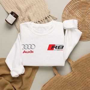 May include: A white crewneck sweatshirt featuring the silver Audi four-ring logo and the red Audi text on the left side, with the Audi Sport R8 logo in red, black, and white on the right side of the chest area.