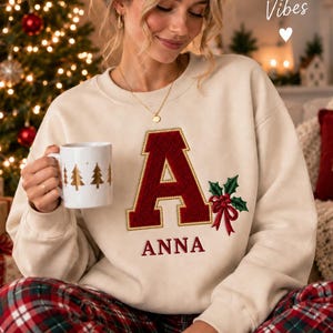 May include: A beige crewneck sweatshirt featuring a large, textured dark red letter A with a gold border, embroidered holly leaves, and a red bow. The name ANNA is embroidered in red below the letter. The text Cozy Christmas Vibes is visible.