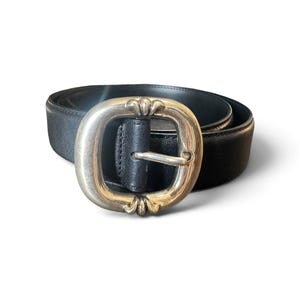 May include: A black leather belt featuring a large, oval-shaped silver-toned metal buckle. The buckle has a smooth, polished finish with decorative, raised floral-like detailing at the top and bottom edges, securing a standard metal prong.
