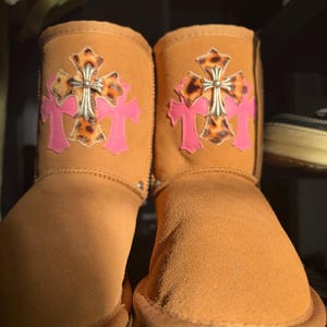 May include: A pair of tan suede ankle boots featuring a decorative patch on the front. The patch consists of a pink cross shape layered behind a leopard print cross, which is topped with a silver-toned metallic cross emblem.