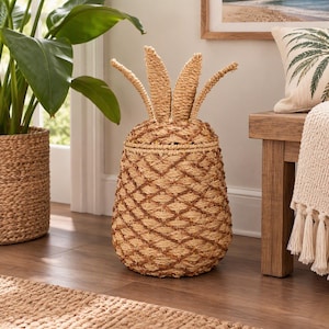 May include: A pineapple-shaped storage basket woven from natural light brown fibers. The basket features a removable lid with upright, pointed leaf-like extensions at the top and a textured, crisscross pattern across the main body.
