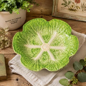 May include: A ceramic bowl shaped like a green cabbage leaf with a scalloped edge and a central white vein pattern. The bowl features a glossy finish with realistic textures mimicking the appearance of a fresh, vibrant green vegetable leaf.