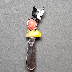 Può includere: Un coltello spalmaburro in acciaio inossidabile con una figura sagomata di Topolino come manico. La figura ha testa nera, pantaloncini rossi e scarpe gialle. La lama in metallo reca l'incisione del testo Disney STAINLESS CHINA sul davanti.
