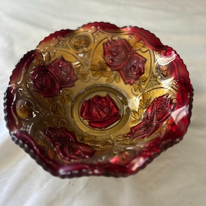 May include: A round glass bowl featuring a deep red and amber gradient finish. The interior surface is decorated with embossed rose motifs and leaves, with a prominent rose centered at the base. The bowl has a scalloped, ruffled edge.
