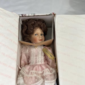 May include: A porcelain doll with curly brown hair and blue eyes, wearing a pink dress with white lace trim and a sheer polka-dot overlay. A gold-colored tag attached to the dress displays the text KRYSTAL #5002. The doll is inside a box.