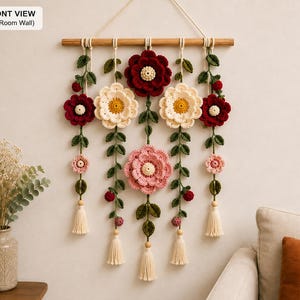 May include: A wall hanging featuring five vertical strands of crocheted flowers in shades of deep red, cream, and soft pink, accented with green leaves and small berries. The strands are suspended from a wooden dowel with cream-colored tassels.