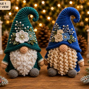 Op de afbeelding: Twee gehaakte kabouters met hoge puntmutsen, één in blauwgroen en één in blauw. Beide mutsen zijn versierd met witte en blauwe bloemen. De blauwgroene kabouter heeft een witte baard met sneeuwvlokpatroon, de blauwe een krullende crèmekleurige baard.
