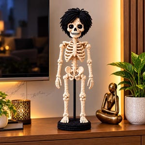 May include: A crocheted skeleton figurine featuring an off-white yarn body with detailed rib cage, pelvic, and limb structures. The figure has a round skull with large black eyes and a thick, textured mop of black yarn hair, standing on a black base.