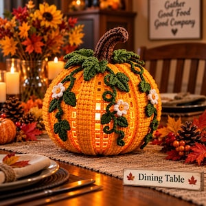 Può includere: Una decorazione a forma di zucca arancione all'uncinetto con un picciolo marrone, foglie verdi all'uncinetto e piccoli fiori bianchi. La zucca ha un corpo cavo a reticolo che si illumina dall'interno e poggia su un tavolo vicino a un cartello Dining Table.
