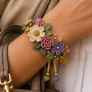 May include: A floral bracelet featuring crocheted flowers in shades of cream, mauve, and purple, accented with green crocheted leaves, small gold-toned beads, and two dangling cords finished with green and purple crocheted spheres and gold beads.