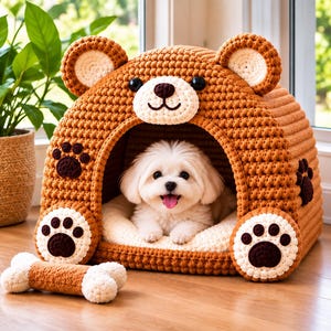 May include: A brown crocheted pet bed shaped like a bear with rounded ears, a snout, and dark eyes. The front features two large white and brown paw prints. A small white and brown crocheted bone toy sits in front of the bed, which houses a small white dog.
