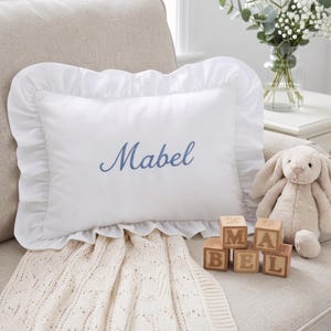 May include: A rectangular white pillow with a ruffled border, featuring the name Mabel embroidered in blue script. The pillow is accompanied by a beige plush rabbit toy and three wooden blocks displaying the letters M, A, B, E, and L.