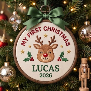 May include: A round wooden embroidery hoop ornament featuring a cream-colored fabric center with an embroidered brown reindeer face, red nose, green holly, and gold stars. The text reads MY FIRST CHRISTMAS, LUCAS, and 2026, topped with a green ribbon.