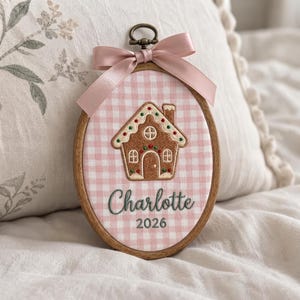 May include: An oval wooden embroidery hoop containing pink and white gingham fabric. The fabric features an embroidered gingerbread house with colorful accents, the name Charlotte in dark green script, and the year 2026 in dark green text.