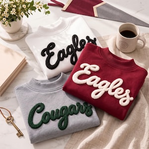 May include: Three crewneck sweatshirts featuring textured, raised lettering. One white sweatshirt with black 'Eagles' text, one maroon sweatshirt with white 'Eagles' text, and one gray sweatshirt with dark green 'Cougars' text. Each garment has a ribbed collar.