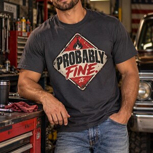 Può includere: Una t-shirt grigio antracite a maniche corte con una grafica a forma di diamante che presenta un'icona a fiamma e il testo "PROBABLY FINE" in grassetto nero e rosso. Il design ha un aspetto usurato ed è abbinato a un jeans blu.