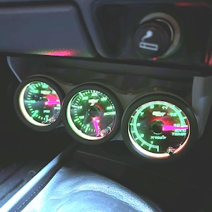 May include: A triple gauge pod mounted in a vehicle dashboard featuring three circular analogue gauges with black faces, green illuminated markings, and bright pink needles. The gauges display PSI, BOOST, and EXT TEMP with clear white text labels.
