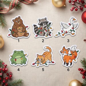 May include: Six embroidered patches featuring cartoon animals tangled in colorful Christmas lights: a brown bear, a raccoon, a white goose, a green frog, an orange tabby cat, and an orange fox, each numbered one through six.
