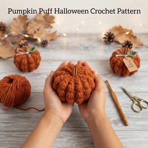 Può includere: Un'intestazione di testo Pumpkin Puff Halloween Crochet Pattern sopra tre zucche all'uncinetto arancioni con steli di bastoncino di cannella, un gomitolo di lana arancione, un uncinetto in legno e forbici dorate su una superficie in legno chiaro.