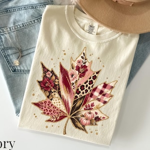 Könnte beinhalten: Ein elfenbeinfarbenes T-Shirt mit einer großen Ahornblatt-Grafik, die aus Blumen-, Leoparden- und Karomustern in Burgunderrot, Rosa und Gold besteht. Das T-Shirt wird mit einer hellblauen Jeans, einem beigen Hut und einer silbernen Kette kombiniert.