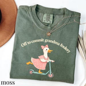 May include: A moss green t-shirt featuring a graphic of a white duck wearing a pink polka dot dress and a matching bow, riding a light blue scooter. The text above the duck reads, Off to commit grandma foolery. The shirt has a crew neckline.