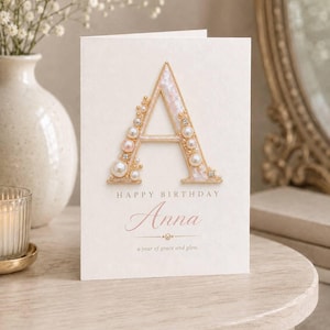 May include: A cream-colored greeting card featuring a large letter A decorated with white and pink pearls and small clear crystals. The text reads HAPPY BIRTHDAY, Anna, and a year of grace and glow. The card has a textured finish and gold accents.