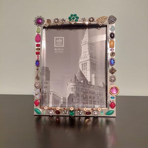 May include: A rectangular silver photo frame measuring 8x10 inches, decorated with an assortment of colorful, multi-shaped faux gemstones and metallic charms along the border. The frame contains a black and white print of a city clock tower.